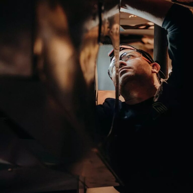 Person wearing safety glasses works on machinery, reaching upward.