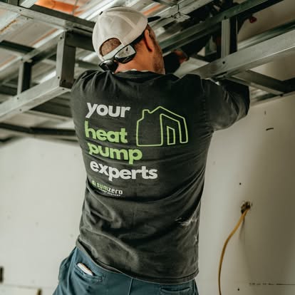 Person wearing a black t-shirt with "your heat pump experts" printed on the back, working under a metal framework.